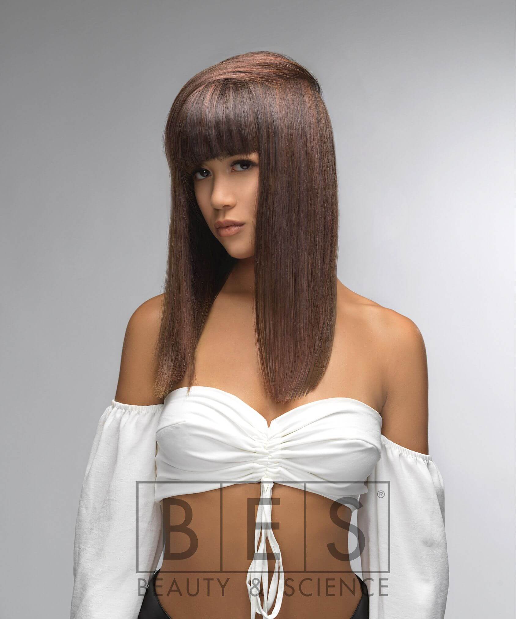 Woman with straight, medium-length brown hair and blunt bangs, wearing a white off-shoulder top, poses against a plain gray background. "BES Beauty & Science" logo is at the bottom.