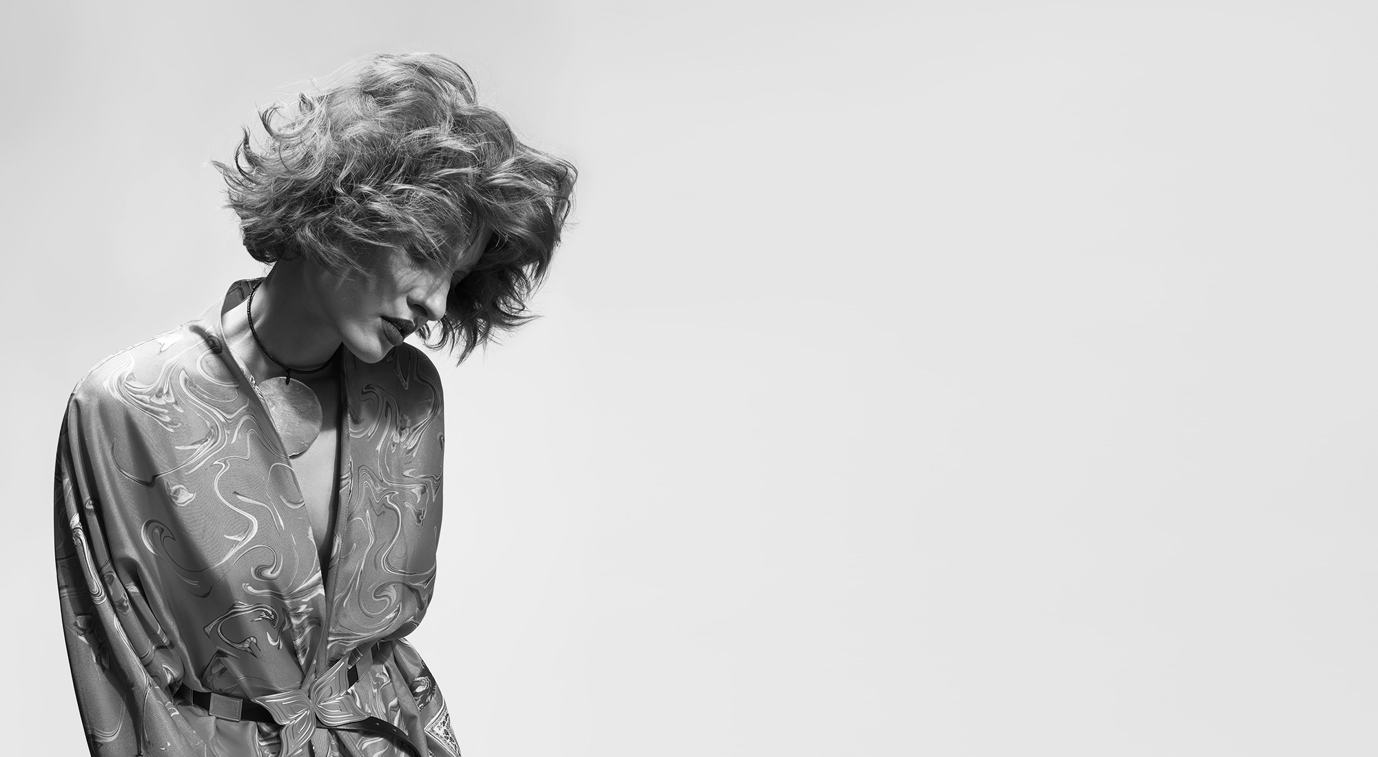 Black and white photo of a person with wavy, tousled hair, wearing a patterned robe, looking down against a plain, light background.